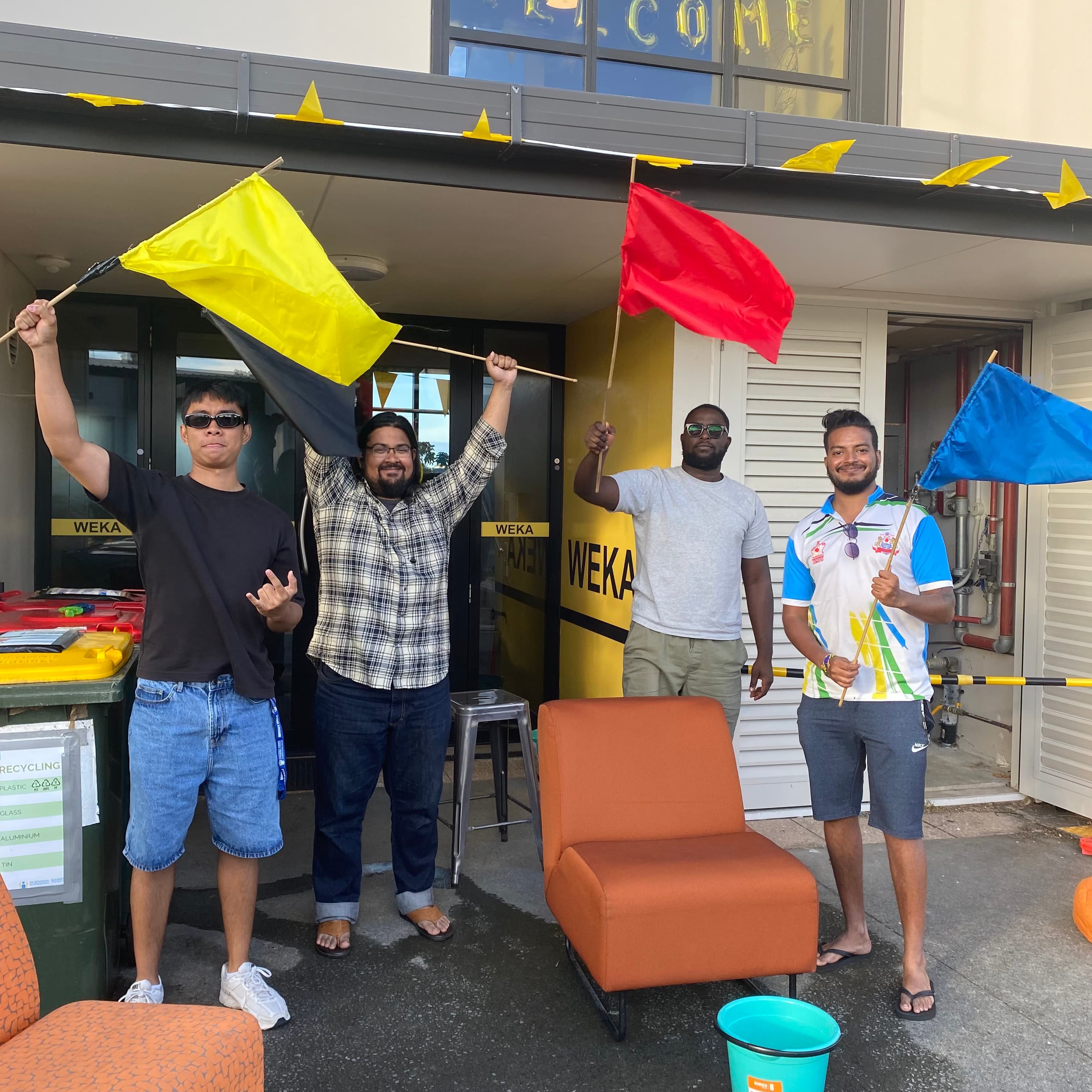 Four Residential Assistants holding team flags outside the Weka block during a village competition.