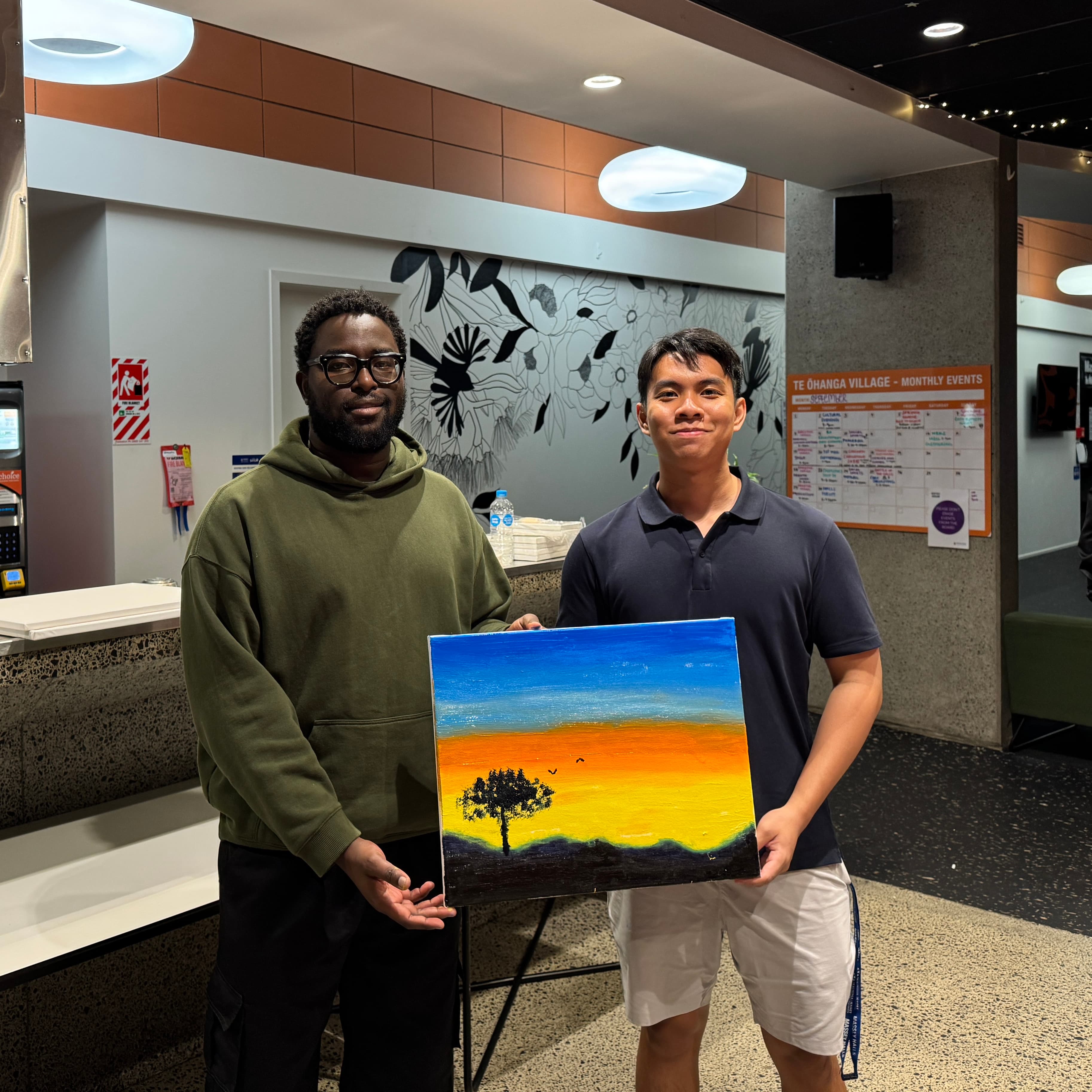 Two residents holding up a sunset landscape canvas from a painting night in the village foyer.